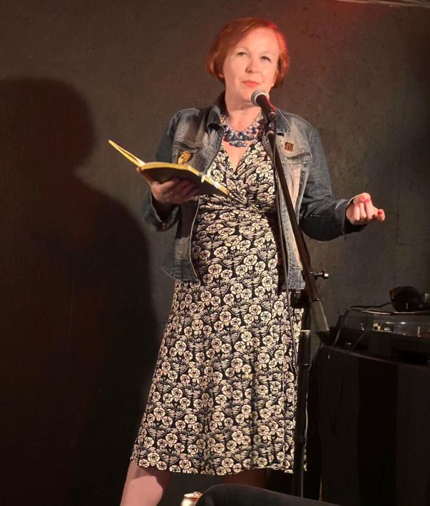 Woman in a dress with red hair holding an open book and talking into a microphone